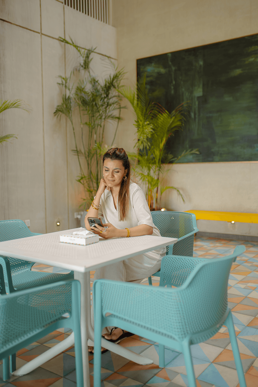 A guest checking her phone at a café table at Cyrus One, surrounded by greenery and artwork in the hotel's shared spaces, Islamabad.