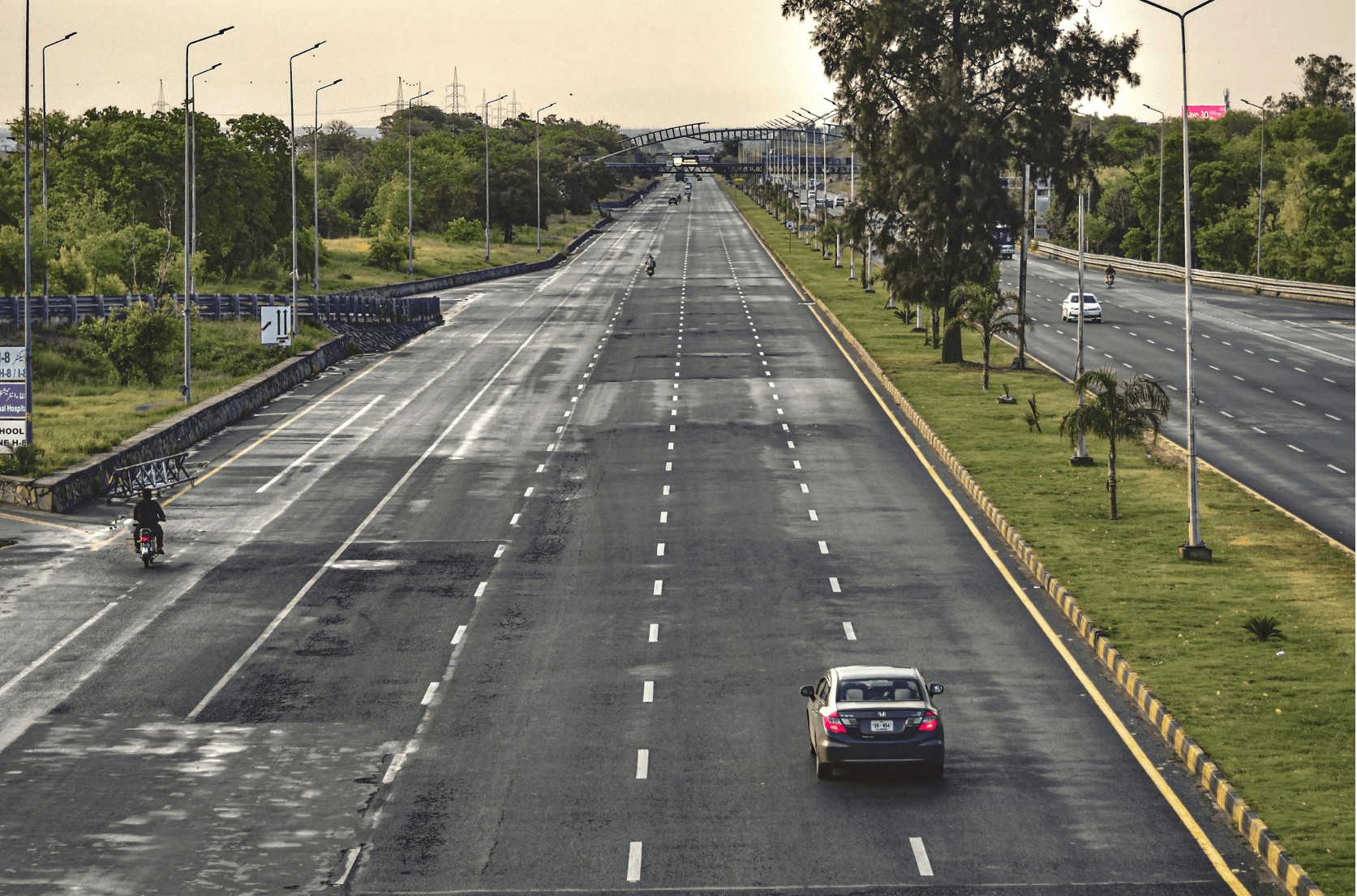 Empty multi-lane highway in Islamabad lined with trees and streetlights, with a motorcyclist and a car in view