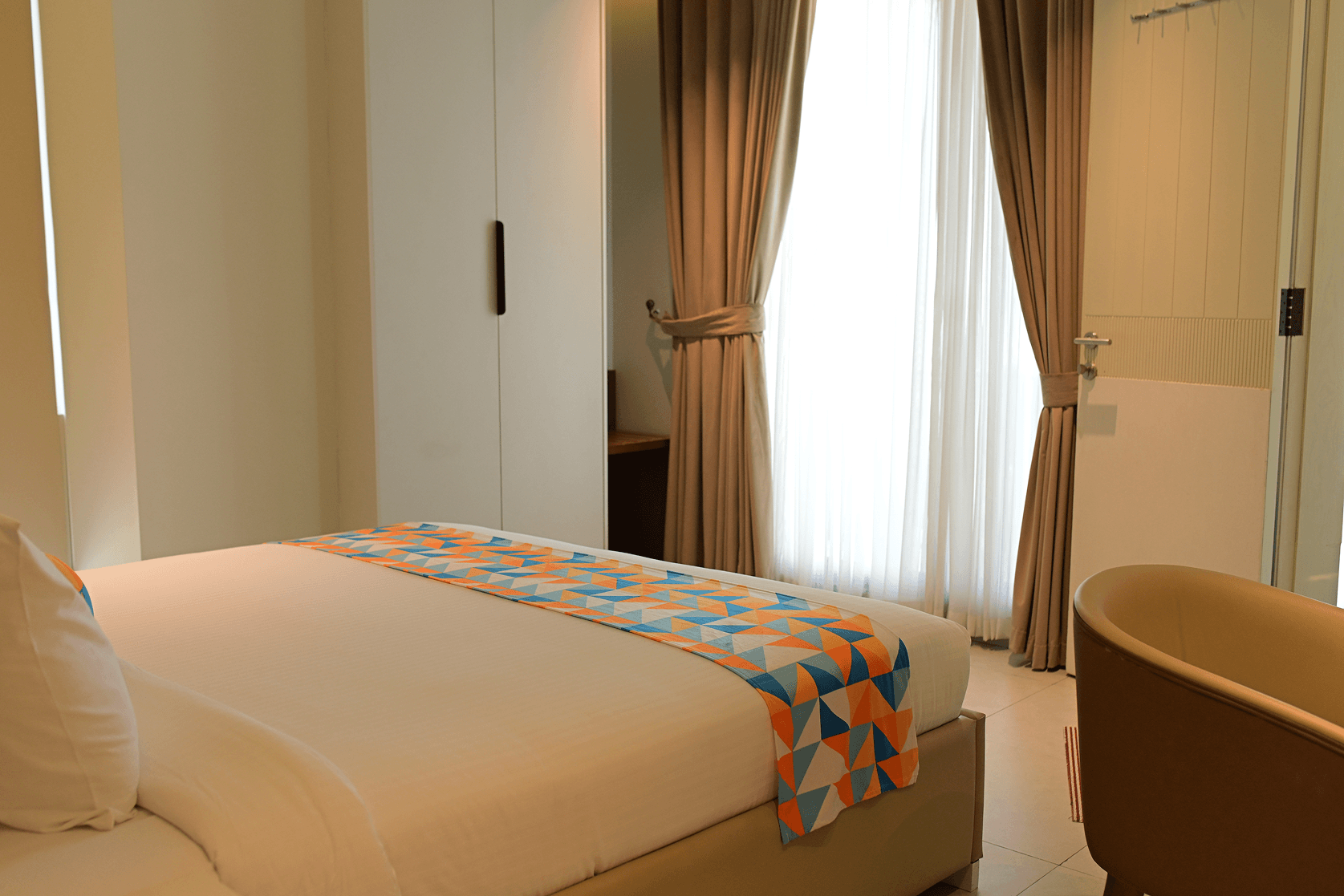 Cyrus One hotel bedroom featuring a crisp white bed with a patterned runner, sheer curtains, and a freestanding tub