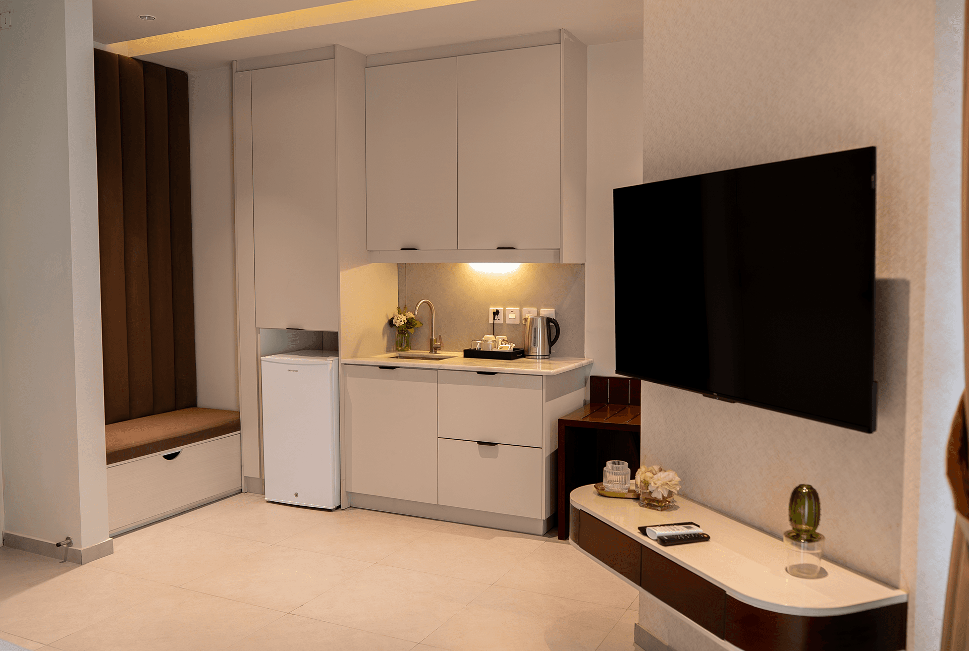 Cyrus One room interior featuring a kitchenette with refrigerator and kettle, seating area, flat-screen TV and comfortable furnishings.